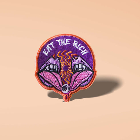 Patch featuring the text 'Eat The Rich' above a design of two open mouths with fangs and a tongue holding an eyeball, set against a purple background with an orange border. The design is bold and edgy.